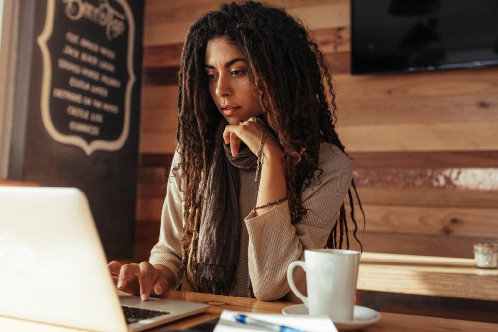 woman sits at table in coffeeshop working on a laptop with a cup of coffee next to her